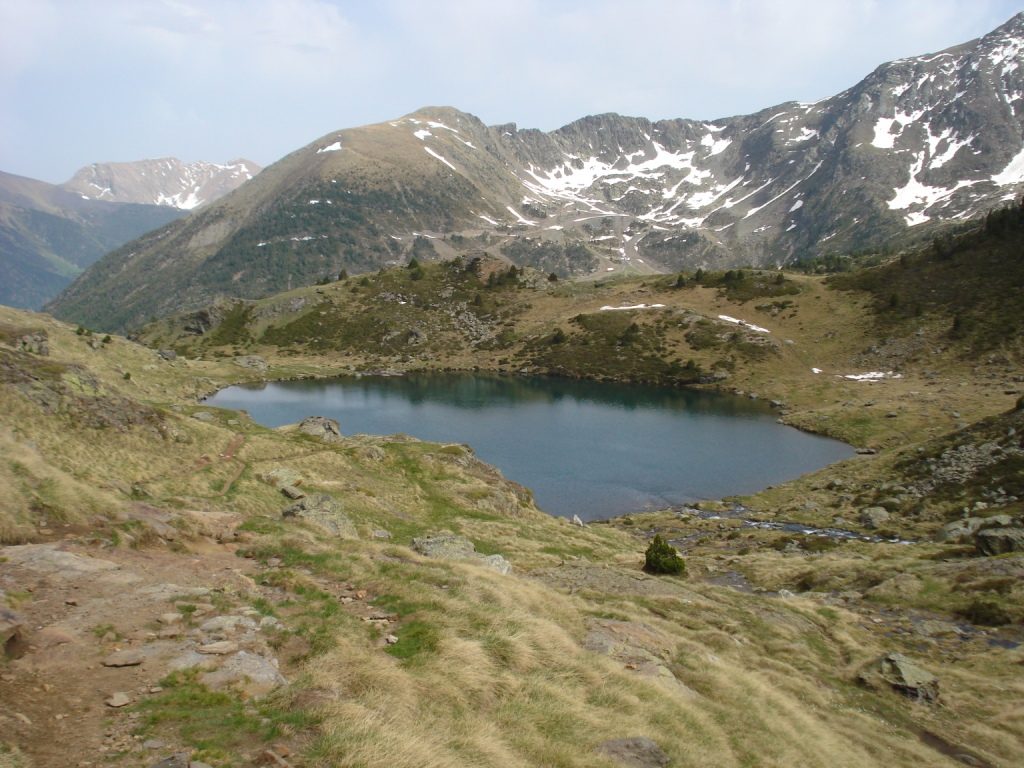 Estany Primer de Tristaina, a deep blue lake in grassy slopes below snow-streaked ridges near Ordino-Arcalis, Andorra